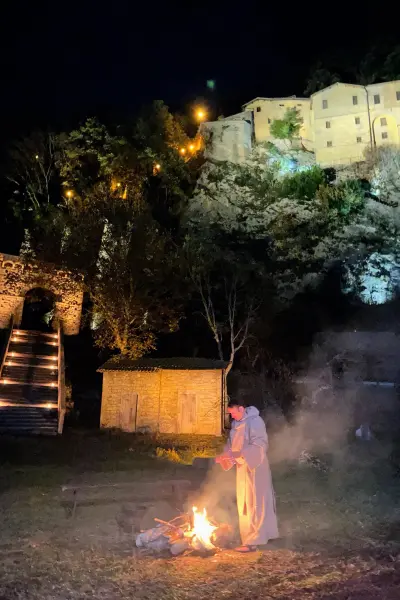 Il Santuario di Greccio illuminato sulla rupe durante il presepe vivente, con un frate accanto al fuoco